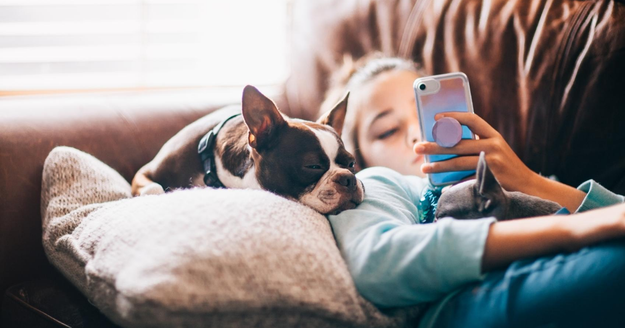 A young teen lays on the sofa with her dog while scrolling on her phone. 