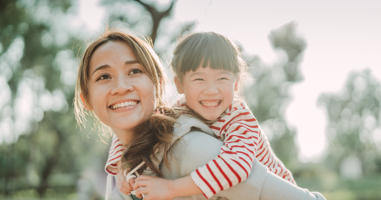 A mother smiles with her young daughter on her back laughing. 