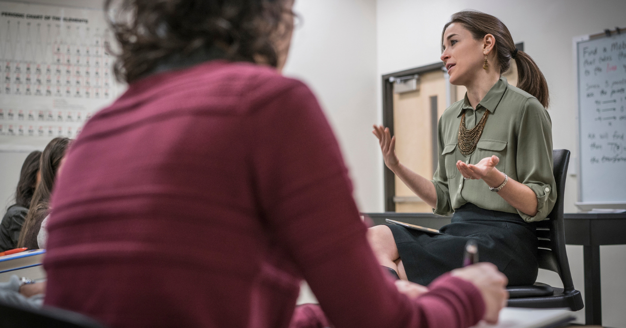 A teacher sits at the front of the classroom and talks to her students as they take notes. 
