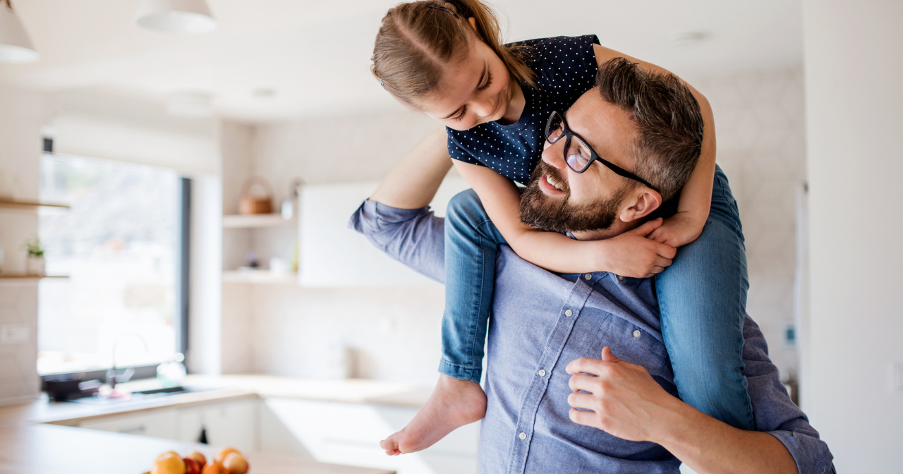 A young girl sits on her father's shoulders inside a house. 