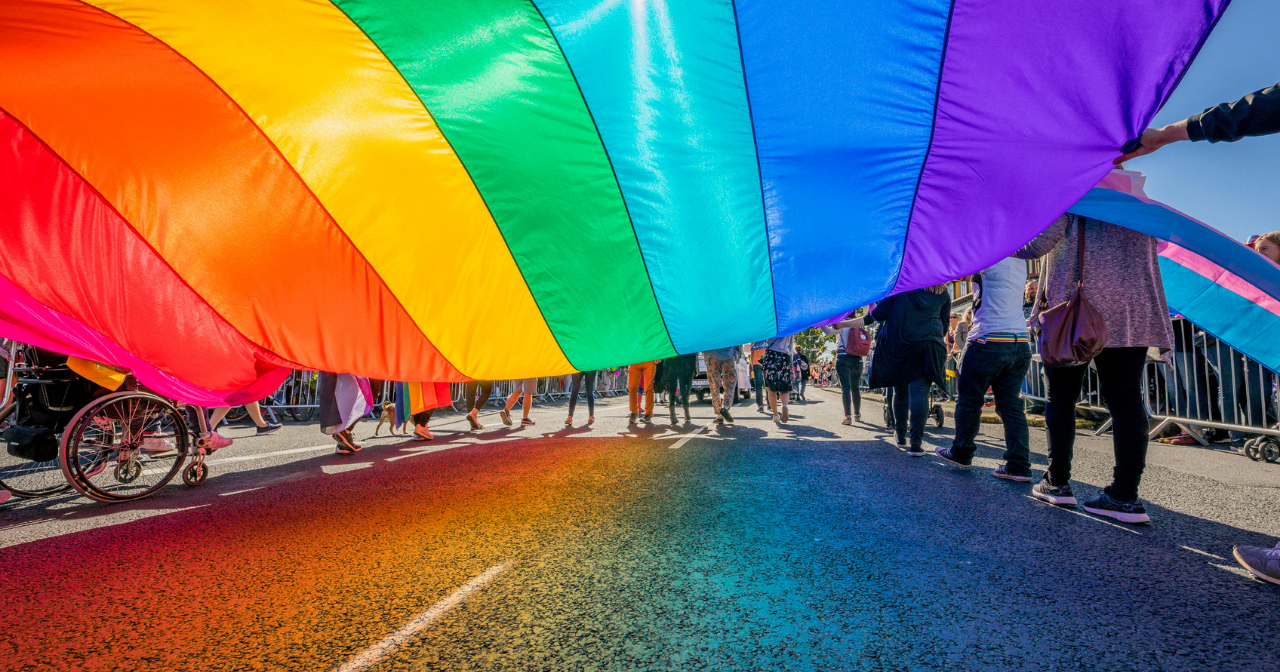 A photo from underneath a massive pride flag being carried by many people during a parade. 