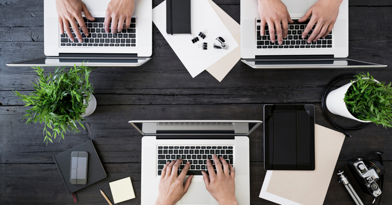 Three sets of hands on three laptops on a large table. 