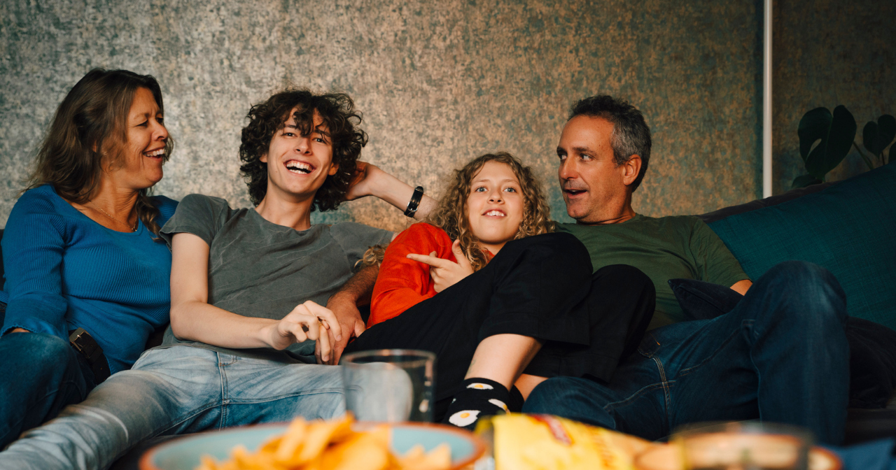 A family of four laugh on the sofa while watching TV. 