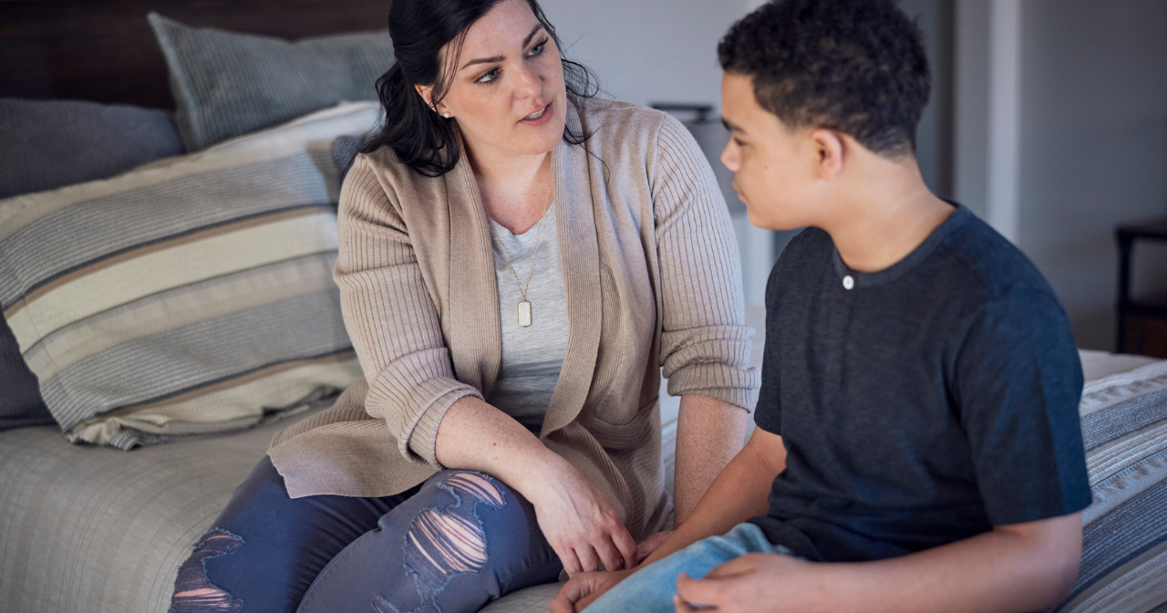 A mom and her son sit on the edge of the bed and talk.