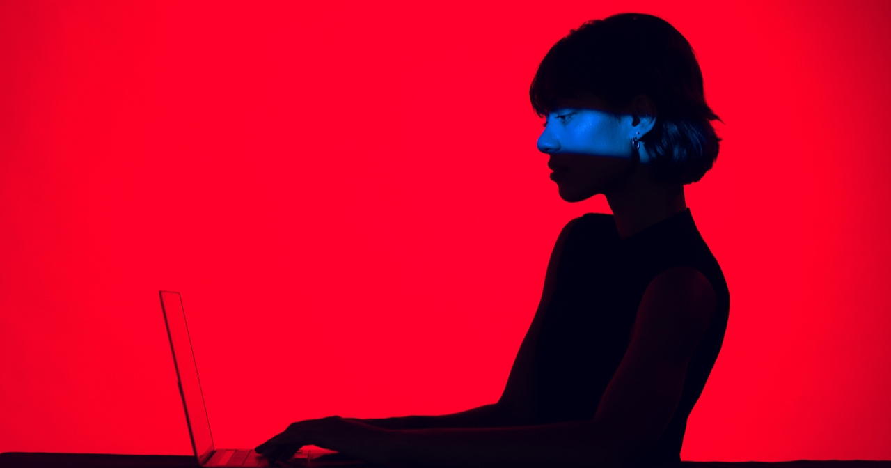 A woman in shadow types on a laptop. The background is bright red.