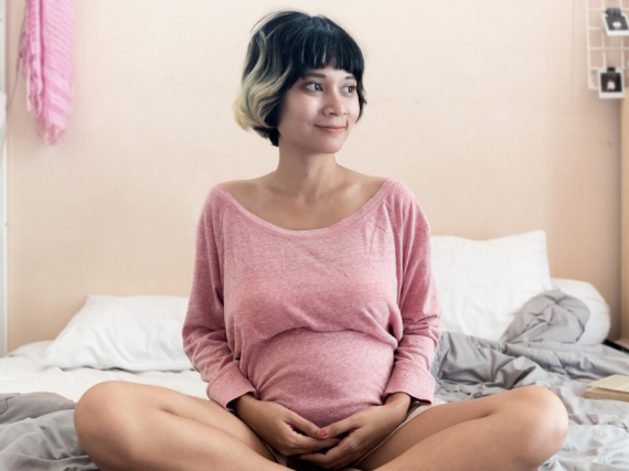 A pregnant woman sits cross legged on a bed