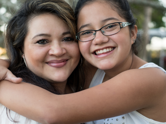 A Latina mom and her daughter hug and smile at the camera. 