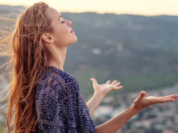 A young woman stands on the top of a hill with her hands outstretched and her face upturned to the sun.