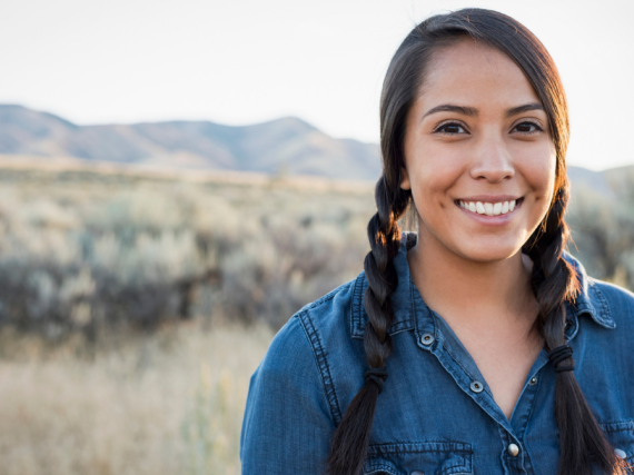A young Native woman smiles for the camera while standing among a field of wild grasses. 