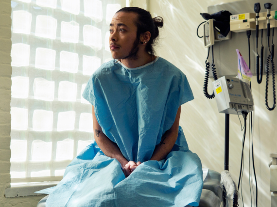 A genderqueer person sits in an exam room wearing a hospital gown. 