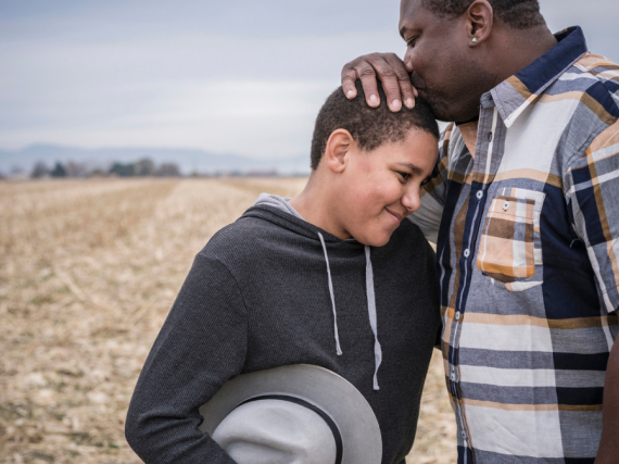 A father and son stand a in a field and the father kisses the top of his son's head. 