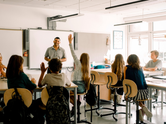 An engaged middle school classroom with a teacher at the head and several hands raised. 