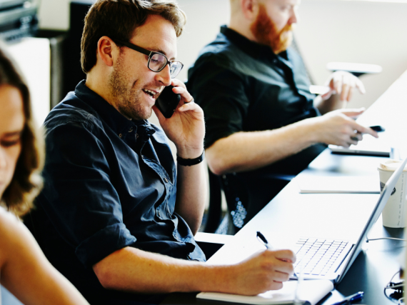Three people sit at their computers while talking on the phone in an office. 