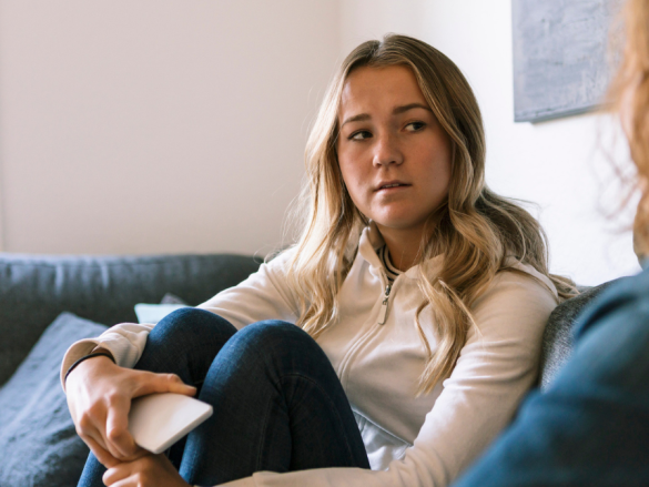 A concerned looking teen hugs her knees to her chest while talking to an adult. 